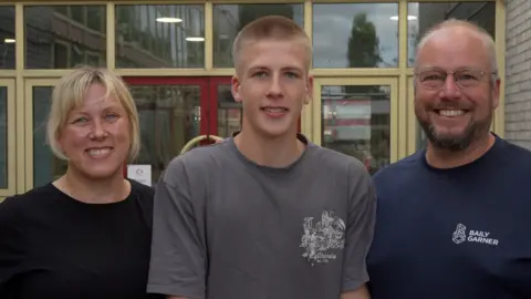 Jamie Niblock/BBC (L-R) A woman with short blonde hair and a fringe smiling at the camera. She is wearing a black top.
A teenager with blonde hair and wearing a grey t-shirt and a man with a big smile wearing clear frame glasses and wearing a navy blue t-shirt.