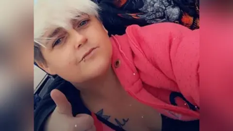 A selfie of a young woman with short blonde hair, wearing a pink hoodie and giving a thumbs up to the camera