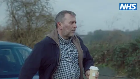 NHS Robert in the NHS video, which shows arms around him, to stimulate how the tightening of a chest can feel like, he is wearing a blue and green checked shirt, he is holding a cup of coffee and is outside by a car. 