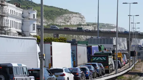 Traffic stationary and queuing along a road near the Port of Dover. Vehicles are also seen on a congested flyover. The white cliffs of Dover can be seen in the background.