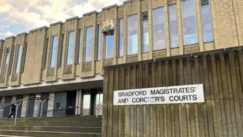 The image shows the exterior of Bradford Magistrates’ and Coroner’s Courts building. The structure is made of concrete with a series of tall, narrow vertical windows across the upper section. Above the windows there is a royal coat of arms. In the foreground there is a set of wide concrete steps leading up to the entrance, which has metal railings on both sides. On the right side of the image, a large vertical wall displays a white rectangular sign with black text that reads: BRADFORD MAGISTRATES’ AND CORONER’S COURTS.