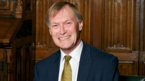 Getty Images Sir David Amess wearing a golden coloured tie and a navy suit. He is standing in front of wood panelling and smiling. 