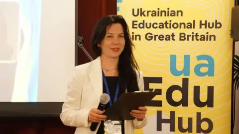 Nataliia Gorbatykh A woman with long brown hair wearing a black dress and white blazer holds a microphone and clipboard. She is smiling. She is standing in front of a screen and a yellow banner that reads: ua Edu Hub