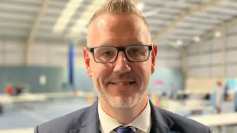 A man wearing a suit jacket is looking ahead at the camera - he has a white suit on with a blue tie. He is wearing glasses.