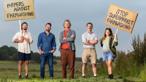 Kriss Woodhead, Jimmy Doherty, Guy Singh Watson, Ben Andrews and Zoe Colville are stood outside in a field with two placards urging supermarkets to top farmwashing