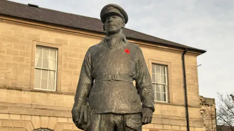 A statue honouring Mayne  - it is a bronze figure with its arms at its side wearing a military uniform including a peaked cap. A red poppy is on its left breast. In the background is a sandstone building.