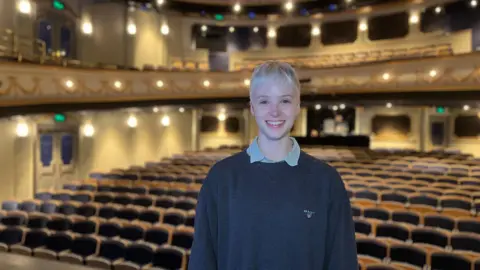 A woman with short blonde hair. She is standing in a theatre. She is wearing a navy crewneck jumper standing in an auditorium. 