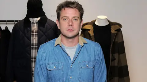 Getty Images Jonathan Anderson looking at the camera, he has dark hair. He is wearing a denim blue jacket with tan collar and a grey shirt