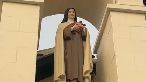 A statue of St Theresa in habit standing in a churches tower holding roses and a crucifix. 