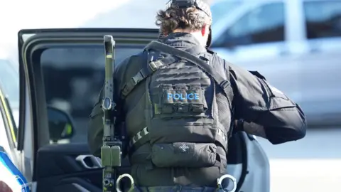 Eddie Mitchell An armed Sussex Police officer during a stop on Woodside Road, Worthing. The officer has his back to the camera and has a gun on his left-hand side. He is exiting a police car, and is wearing a bullet proof vest.