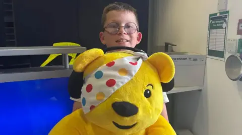 A boy with brown hair and glasses behind a large yellow teddy bear with a spotted cloth over one eye