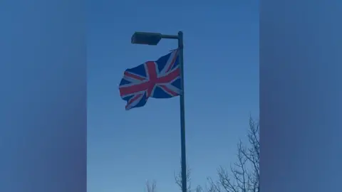 Bristol Rises A Union flag is tied to the top of a lamppost in shaded area outside a hospital. There are naked trees in the background and a brown building.