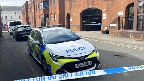 A police car parked up on Bedford Street behind a blue and white cordon. In the background you can see an arched entrance into a brick building where a cordon has been placed.