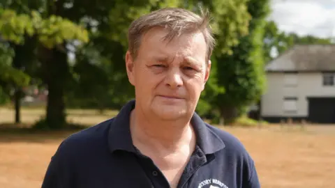 Shaun Whitmore/BBC A man with short brown hair looks directly at the camera in a head and shoulders image. He is wearing a navy polo shirt and standing on a village green, with trees behind him and a white house