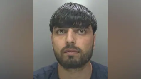 Bilal Dawlatzai in a police custody shot looking at the camera. He has dark hair falling over his eyebrows and facial hair. He is wearing a dark blue T-shirt.