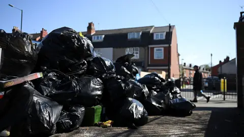 Reuters A big pile of black bin bags on a street on 2 April. A man in background walks past, with houses behind the bin bags