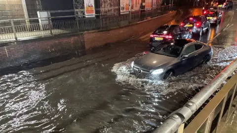 A flooded road in Cardiff city centre. A silver Mercedes attempts to drive through the water. 