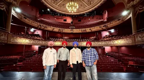 Wolverhampton Grand Rangle Sardar on the stage at the theatre. The four men stand in a line with the seats of the theatre behind them. They are all wearing shirts, jeans or black trousers and turbans. 