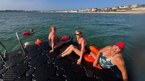 Helene Frankham Three women sit on a raft in the sea with Greenhill beach in the background