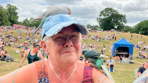 Ollie Ward/BBC A woman wearing a denim cap and pink and orange dress smiles at the camera. There is a small white bird perched on her cap while a small green bird rests on her shoulder.
