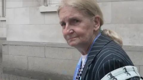 An elderly woman with tied-back blonde hair wearing a striped blazer.