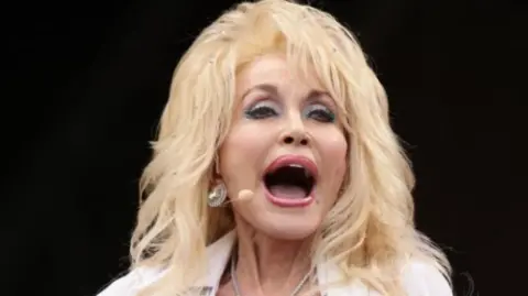 PA Media Dolly Parton, wearing all white, plays on the Pyramid Stage at the Glastonbury Festival in 2014