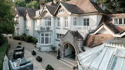 A view of the exterior of Chateau Le Chaire. It is a large white building with many windows. There's a conservatory to the right, and garden furniture to the right.
