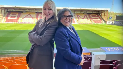 BBC/Aisha Iqbal Two ladies pose back to back in the stands of a football stadium. They are both smartly dressed in suits. One is younger (maybe in her 30s) and is white, with long blond hair. She wears a grey suit. The other lady is middle aged, and of South Asian heritage. She wears a blue suit and black glasses. It is a clear sunny day, and the green, lush pitch is visible behind them along with a stand with seating spelling out 'Bantams'.