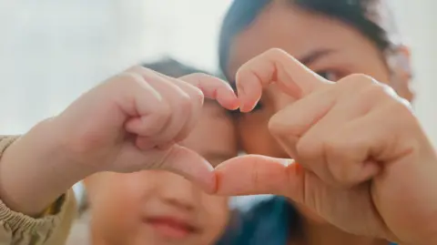 A stock image of two people making a love heart symbol with their hands. It is close up of the hands but in the background you can see a young boy and an older woman. 