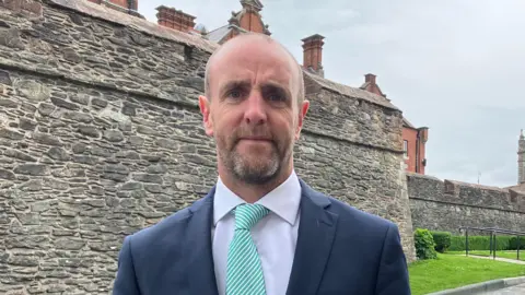 Mark H Durkan is wearing a green and white striped tie, white shirt, and a navy blazer. He is standing near to Derry's historic walls.