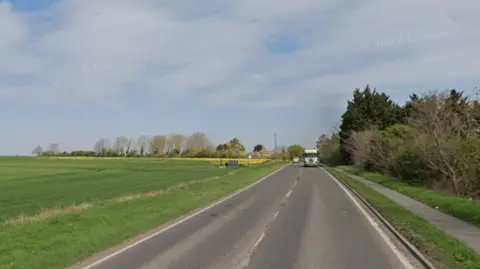 Google image of the A10 during the day. There is grass on either side of the road and a flat field. 