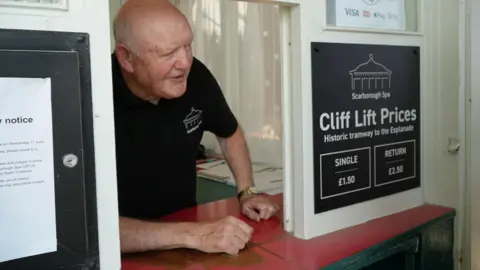North Yorkshire Council An older man leans out of a red and white ticket kiosk window. He wears a black polo shirt with a white Scarborough Spa logo on. To the left, a black board displays the Scarborough Spa logo and ticket prices.