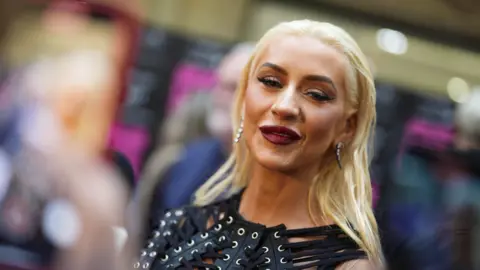 A woman with blonde her combed back. She is wearing red lipstick and a black outfit. She has pearl hoop earrings and looking at the camera 
