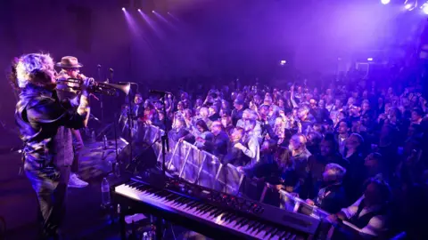 @Slow Moving Media A performer at the Cheltenham Jazz Festival sings for the crowd under purple lighting on the main stage at the Cheltenham Jazz Festival.