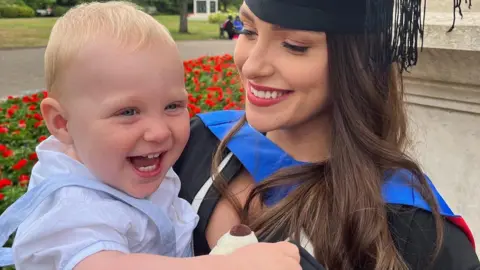 Emma Powell Leo with a big smile in his mums arms. She is looking at him smiling. She is wearning a graduation cap and gown. He is wearing a light shift and trousers with suspenders. He has wispy blonde hair