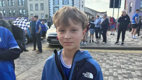 James. He has light brown hair, wearing a blue North Face jacket and blue Swifts jersey underneath. Other people are in the background.