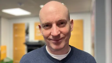 A man is smiling at the camera he is largely bald. He is wearing a blue crew neck jumper above a white t-shirt 