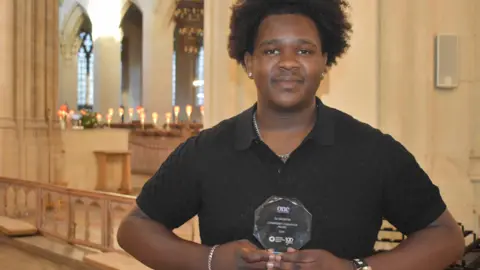 Suffolk One Sixth Form College DJ Mudziviri, who is wearing a black t-shirt, holding a small glass trophy. He is standing in what looks like a church setting. He is looking directly into the camera.