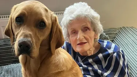 Hannah Kent A golden-brown dog sits upright on a gray cushioned sofa in front of an older person. The dog has floppy ears and a smooth coat. The person, Joan, wears a blue-and-white striped sweater.