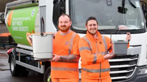 Two men in orange jump suits with Chichester District Council logos on them. They are smiling and holding small bins. They are standing in front of a lorry.