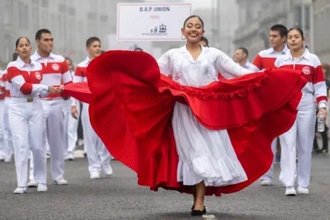 Thomas Andy Branson A group in white and red outfits carrying a banner reading "BAP Union Peru" with a dancer in a white dress and flowing red skirt dances ahead, parading down the street