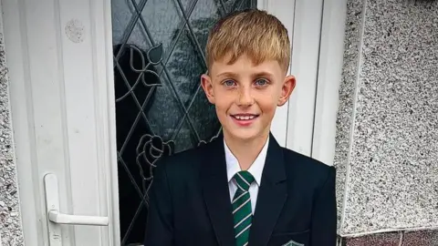 Handout A boy with blonde hair wearing a school uniform which comprises of a green and white striped tie and a blazer. 