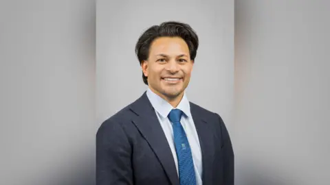 Cambridge University Hospitals NHS Foundation Trust Photo of Dr Nuru Noor against a light blue background. He is smiling and wearing a dark blue jacket, light blue shirt and tie. 