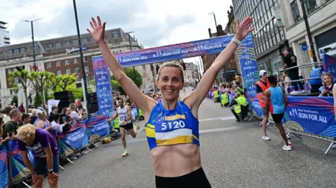 A woman stands at the finish line of the Leeds 10 with her arms in the air. She has brown hair and is smiling. She is wearing a blue sports bra.