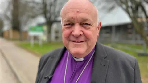 A smiling bishop with very short grey hair on the sides of his head, in a purple shirt, dog collar and grey jacket stands in front of some trees and a strip of grass.