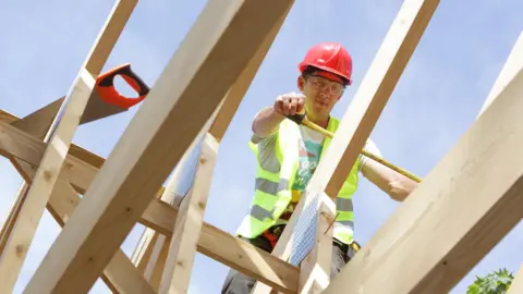 A carpenter wearing hi vis and a hard hat, works on the roof beams of a house under construction. A saw with an orange handle sits on one of the beams.