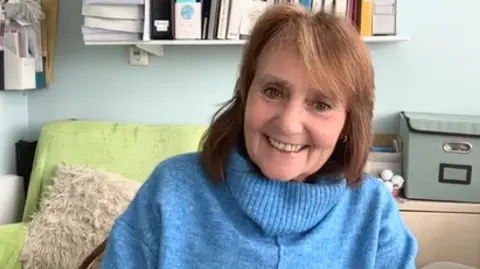 Judith Cantrell has brown hair with a fringe and is wearing a pale blue jumper. She is sitting at a desk and behind her there are books on shelves, a pale lime-green sofa and a moss green storage box. She is smiling.
