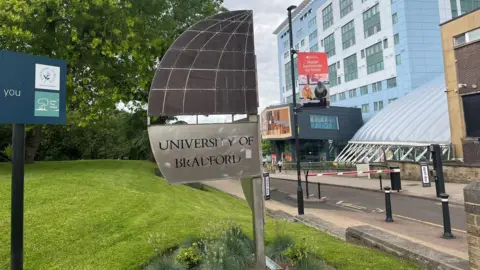 Les images BBC / Adam Laver montrent l'entrée de l'Université Bradford. Au premier plan, il y a un signe métal qui affiche clairement le nom de l'université. "Université de Bradford". Ce panneau est situé dans des zones herbeuses faites de plusieurs plantes. Un grand bâtiment moderne peut être vu en arrière-plan. Le bâtiment a un extérieur bleu et blanc, lui donnant un look moderne. À gauche de l'image se trouve un panneau noir avec du texte vert et blanc, et un autre panneau rouge près du bâtiment.