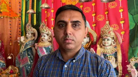 Jadzia Samuel / BBC A man wearing a blue checked shirt. He is stood in front of brightly coloured wall hangings and small statues of people with Hindu clothing on.