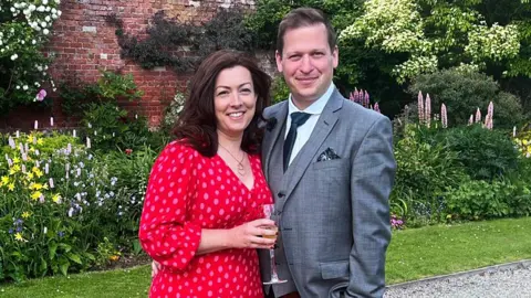 Ben Howard Ben and Sarah Howard standing in a garden. Sarah is wearing a red dress with an all-over spot pattern and holding a drink. Ben is wearing a grey suit and dark tie